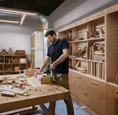 A person wearing safety equipment uses a cordless drill on a wooden box in a workshop.