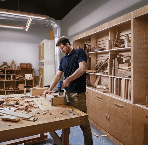 A person wearing safety equipment drills into wood on a workbench in a carpentry shop.