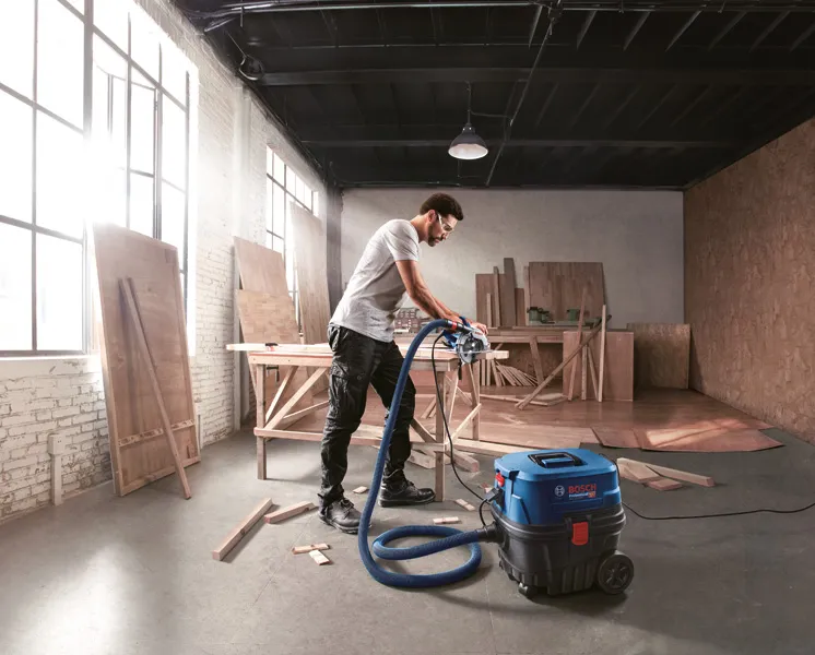 GAS 12-25 PL A person wearing safety equipment uses a vacuum in a wood workshop.