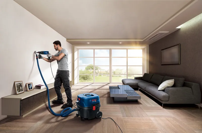 GAS 12-25 PL A person drills into a living room wall using a power tool connected to a dust extractor.