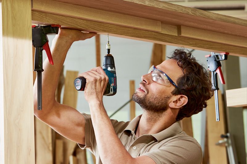 GSR 12V-35 A person wearing safety equipment uses a cordless drill to fasten a screw into wooden beams.