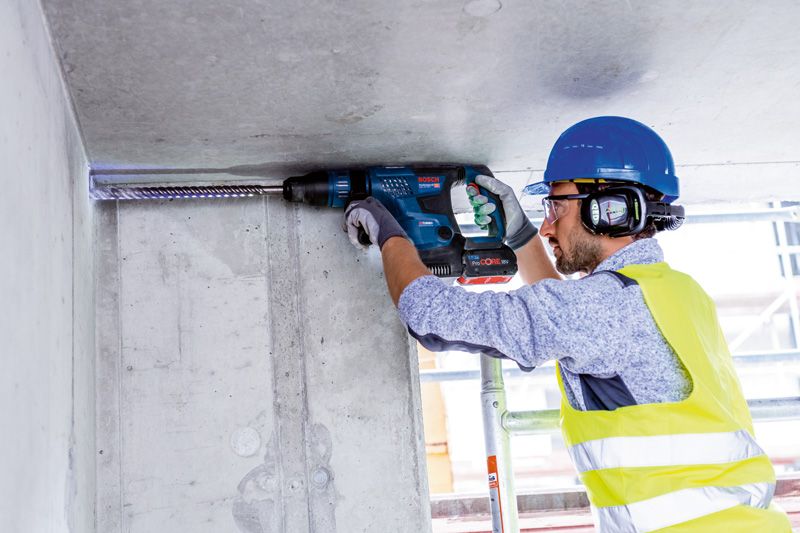 A person wearing safety equipment drills into a concrete ceiling with a rotary hammer.