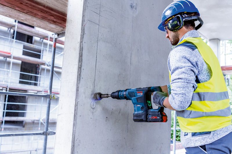 Person wearing safety equipment drills into a concrete wall with a cordless rotary hammer.
