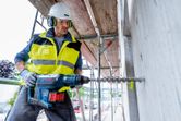 A person wearing safety equipment drills into concrete with a cordless rotary hammer.