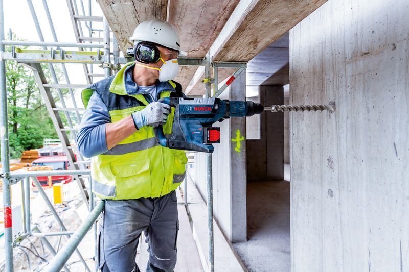 A person wearing safety equipment uses a cordless rotary hammer to drill into concrete.