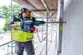 A person wearing safety equipment drills into a concrete wall at a construction site.