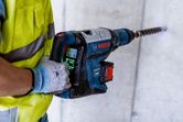 A person wearing safety equipment drills into concrete with a large rotary hammer.
