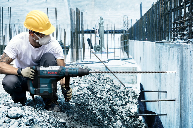 Person wearing safety equipment drills into concrete at a construction site.