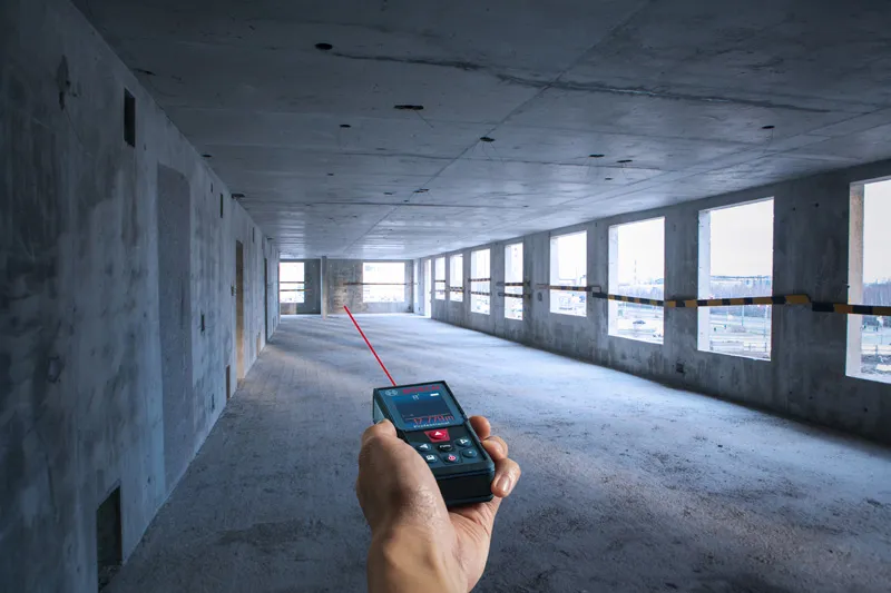 Person using a laser measure to determine distance in an unfinished concrete room.