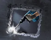 A person wearing safety equipment uses a demolition hammer to break a concrete wall.