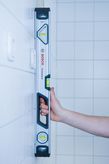 Person holding a 60cm optical level upright against a tiled wall.