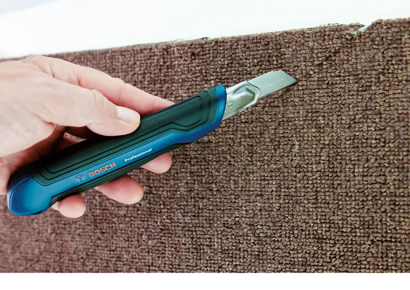 A person cuts carpet along the edge using a utility knife.