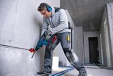 A person wearing safety equipment drills into concrete with a cordless rotary hammer.