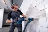 A person wearing safety equipment removes wall tiles using a cordless rotary hammer.