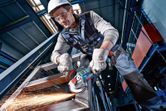 A person wearing safety equipment grinds a metal beam with a power tool, producing sparks.