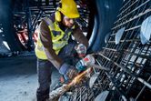 A person wearing safety equipment uses a cordless angle grinder to cut rebar.