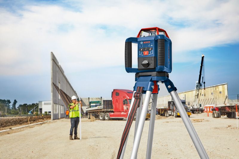 Laser leveling tool on tripod at a construction site, person wearing safety equipment in background.