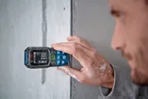 Person measures a wall with a laser measure, paint visible on their hand.