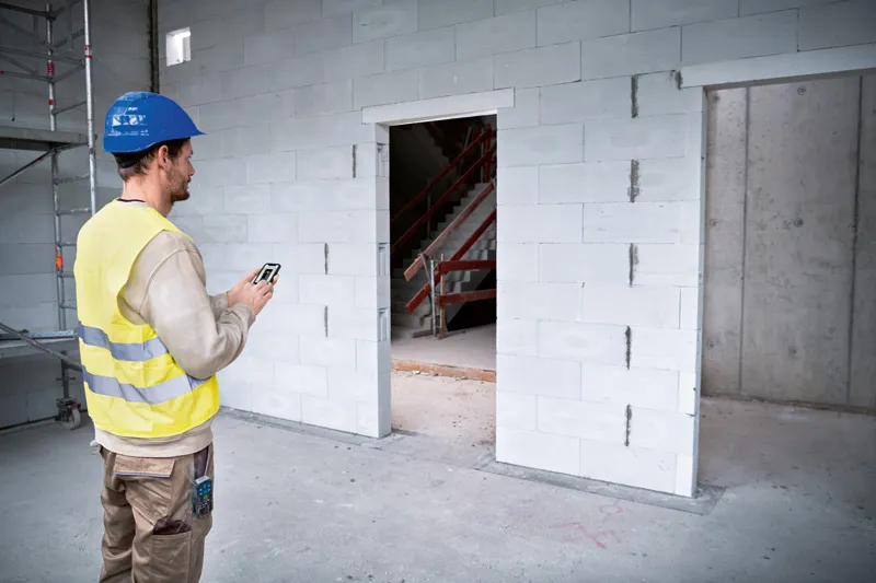 Person wearing safety equipment uses a laser measure to check doorway dimensions on a construction site.