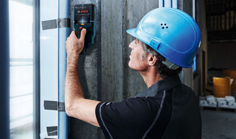A person wearing safety equipment scans a concrete wall with a detector tool.