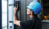 A person wearing safety equipment scans a concrete wall with a detector tool.