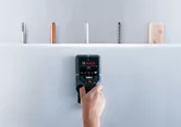 A person scans a drywall with a detector, identifying hidden pipes and wires.
