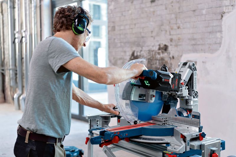 A person wearing safety equipment operates a cordless mitre saw on a workbench.