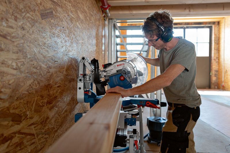 A person wearing safety equipment cuts a wooden beam with a cordless mitre saw.