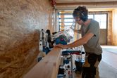 A person wearing safety equipment cuts a wooden beam with a cordless mitre saw.