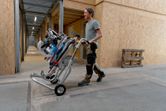 A person wearing safety equipment moves a cordless mitre saw on a wheeled stand indoors.