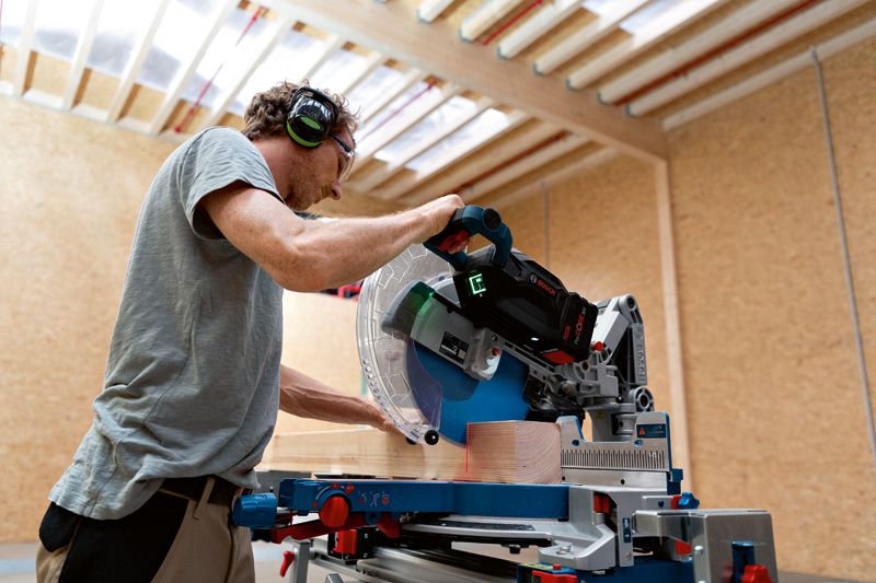 A person wearing safety equipment uses a cordless mitre saw to cut wood.