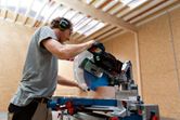 A person wearing safety equipment uses a cordless mitre saw to cut wood indoors.