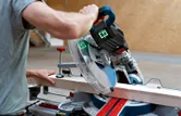 Person operates a cordless mitre saw to cut a wooden plank on a workshop table.