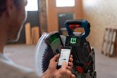 A person adjusts a cordless mitre saw with a smartphone in an indoor workshop.