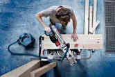 A person wearing safety equipment cuts a wooden plank with a cordless mitre saw.