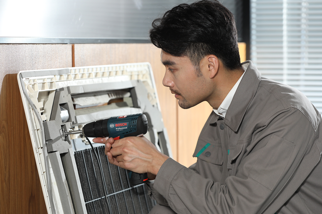 Person wearing safety equipment uses a cordless drill on an air conditioning unit.