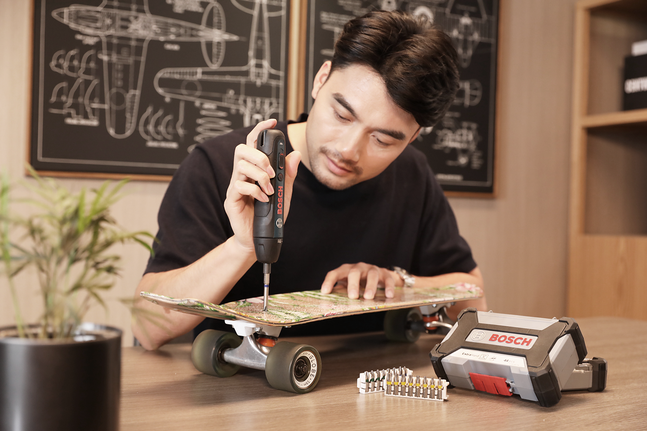 Person uses a cordless screwdriver to repair a skateboard on a desk.