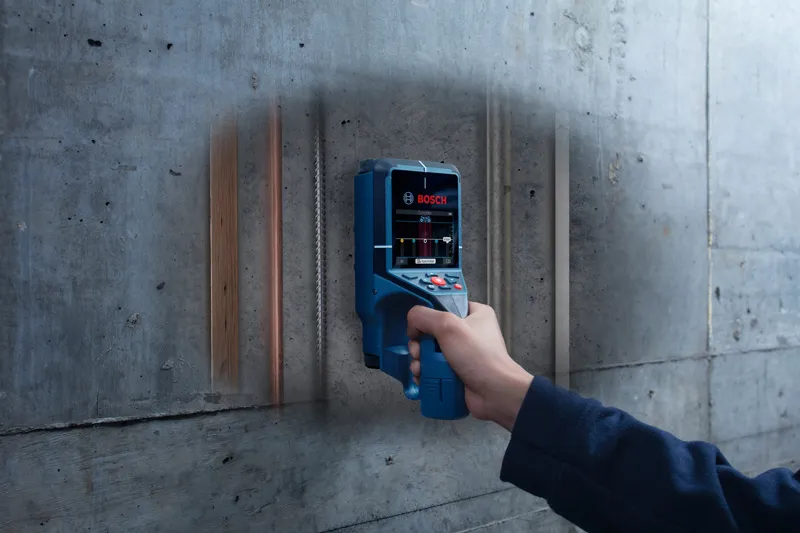 Person scanning a concrete wall with a digital detector, revealing pipes and wiring inside.
