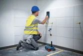 A person wearing safety equipment uses a wall scanner on tiled wall.
