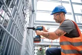 A person wearing safety equipment uses a cordless impact wrench on a metal fence.