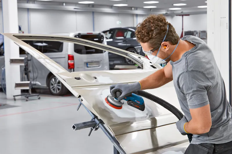 A person wearing safety equipment sands a car door with a power sander in an auto workshop.