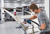 A person wearing safety equipment sands a car door with a power sander in an auto workshop.