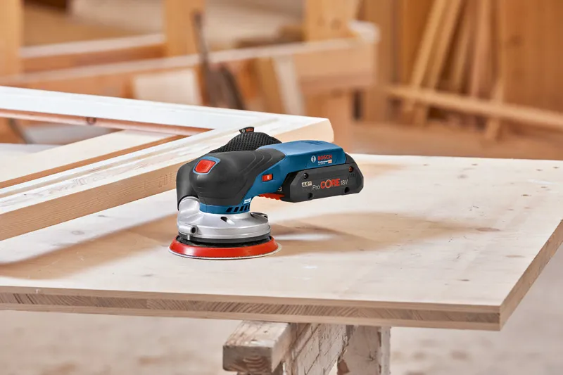Cordless orbital sander resting on a wooden board in a carpentry workshop.