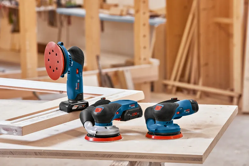 Three electric sanders rest on a wooden workbench in a carpentry workshop.
