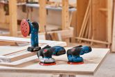 Three electric sanders rest on a wooden workbench in a carpentry workshop.