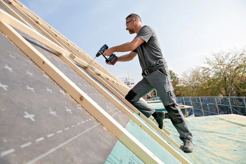 GSR 18V-150 C A person wearing safety equipment uses a cordless drill to fasten wood on a sloped roof.