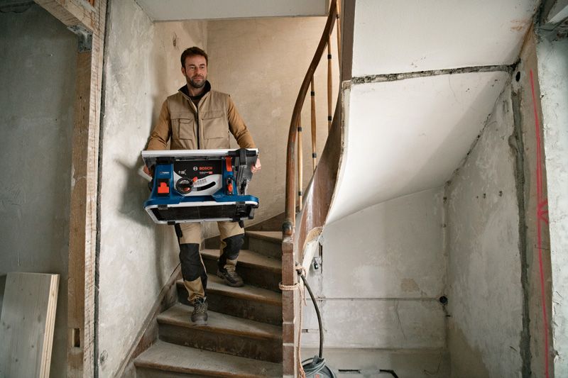 A person wearing safety equipment carries a cordless table saw down a staircase.