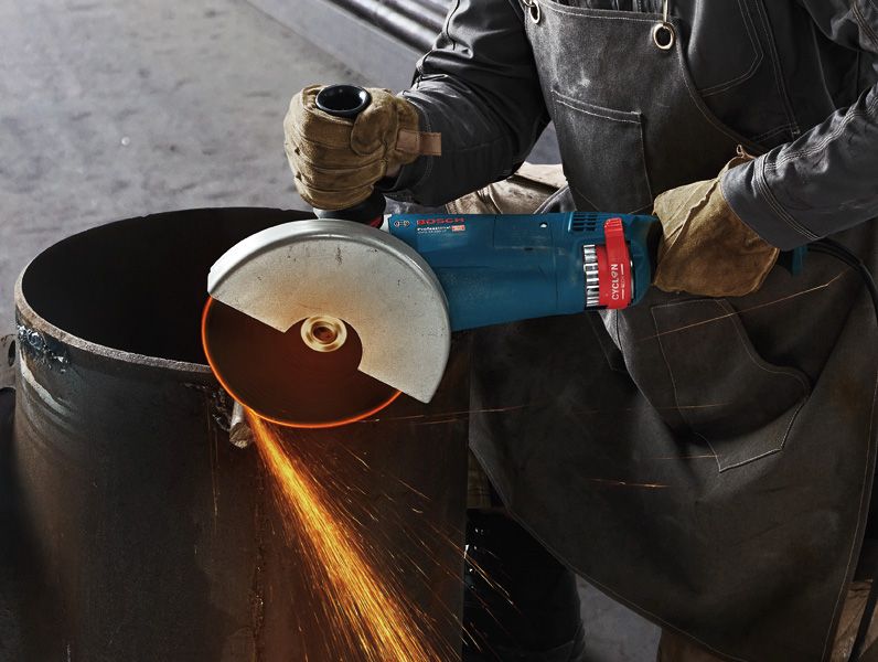 A person wearing safety equipment cuts a metal pipe with an angle grinder.