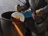 A person wearing safety equipment cuts a metal pipe with an angle grinder.