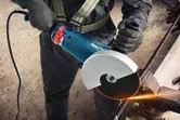 A person wearing safety equipment cuts metal rebar with an angle grinder, creating sparks.